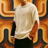 Men’s cream cable-knit sweater featuring contrasting sleeves in black and rust orange, photographed in front of a bold vintage geometric wallpaper in brown, orange, and navy hues.