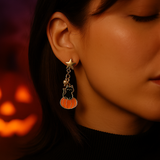 Close-up of a person wearing a Halloween-themed earring with a pumpkin and cat design, with a blurred pumpkin in the background.