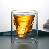 Double-walled glass with a skull-shaped inner design filled with amber liquor, spooky Halloween drinking glass.