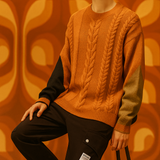 Men’s burnt orange cable-knit sweater with contrasting black and beige sleeves, paired with dark trousers, set against a retro 70s orange patterned background.