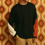Men’s black cable-knit crew sweater with dual-tone sleeves in ivory and burnt orange, styled against a 1970s geometric brown and orange wallpaper backdrop.