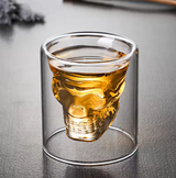 Double-walled glass with a skull-shaped inner design filled with amber liquor, spooky Halloween drinking glass.