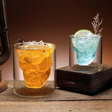 Double-walled glasses with a skull-shaped inner design filled with amber and blue liquor, spooky Halloween drinking glass.